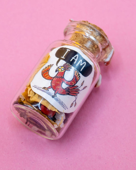 A small Happiness Enchanters handmade glass spell bottle labelled “I AM” featuring a red cartoon phoenix illustration by Katie Abey. The bottle is filled with dried herbs and crystals and sealed with wax. Photographed on a pink background. A magical affirmation gift available at Rebel Romance, a UK brand celebrating alternative style and mindful magic.