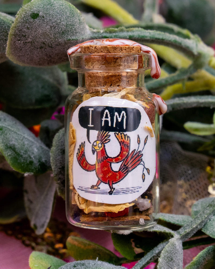 A mini spell potion bottle available at Rebel Romance featuring a red phoenix with the words “I AM” illustrated by Katie Abey. Surrounded by faux leaves, the bottle contains dried herbs and crystals, handmade under the Libra full moon. A magical affirmation bottle from Happiness Enchanters, created by Katie Abey and Angela Sandland.