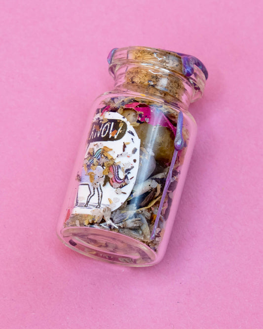 Flat lay of the Happiness Enchanters “I Know” Unicorn Spell Bottle on a pink surface. The 4cm glass bottle features a pastel unicorn label with the words “I KNOW” and is filled with herbs and crystals. A sealed cork top with purple wax adds a magical touch. Perfect for alternative self-care and ritual practices.