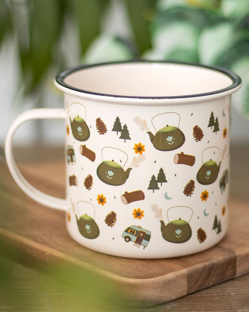 Cream enamel camping mug with a black rim, decorated with a cosy illustrated pattern of green kettles, pine trees, pinecones, logs, yellow flowers, crescent moons, and camper vans, resting on a wooden surface with blurred greenery in the background.