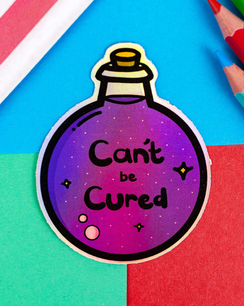 A holographic vinyl sticker by Rebel Romance shaped like a purple potion bottle with a cork stopper, illustrated by Innabox. The label reads “Can’t be Cured” in black lettering on a sparkling background with stars and bubbles. The sticker is placed on a colourful red, green, and blue paper background beside coloured pencils.