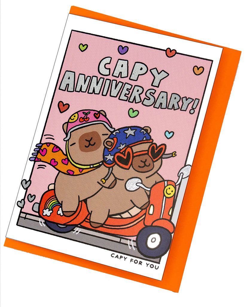Cute anniversary card from Rebel Romance featuring two capybaras riding a red scooter, dressed in fun helmets, heart sunglasses, and scarves. Surrounded by rainbow hearts on a pink background with shiny silver foil text that reads “Capy Anniversary!” Perfect for celebrating love with a quirky, alternative twist. Includes a bright orange envelope.