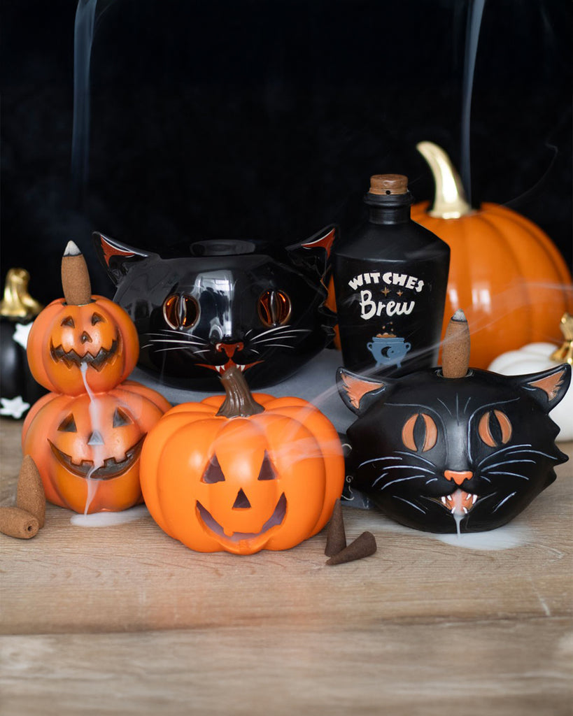 A spooky Halloween-themed setup featuring a black cat ceramic oil burner from Rebel Romance. Surrounded by pumpkin-shaped incense burners, a "Witches' Brew" bottle, and spooky decor, with fragrant smoke rising from the scene. Perfect for alternative home aesthetics.