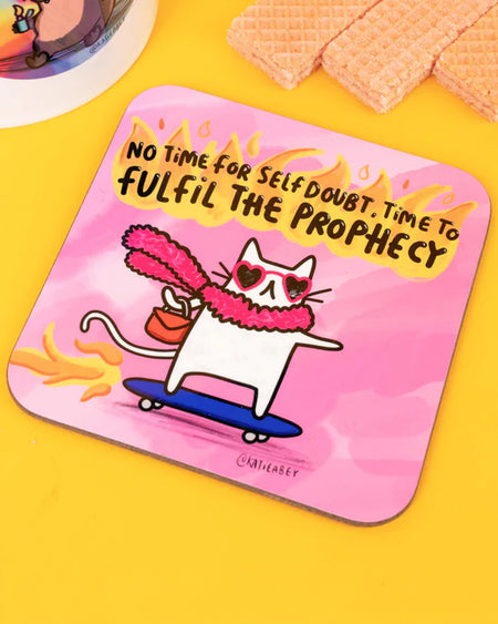 A pink coaster featuring a confident white cat wearing heart-shaped sunglasses and a fluffy scarf, riding a blue skateboard with flames coming from behind. The cat holds a red handbag and is surrounded by the words “No time for self doubt, time to fulfil the prophecy” in bold black and yellow text. Photographed on a bright yellow background with wafers and part of a katie abey moo deng mug visible.