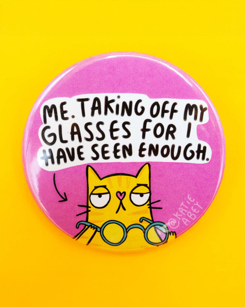 Pink circular badge with a cartoon yellow cat pulling off their glasses in exasperation, alongside the quote "Me. Taking off my glasses for I have seen enough." Designed by Katie Abey for Rebel Romance, a UK small business that celebrates alternative aesthetics. Badge is set against a bold yellow background.