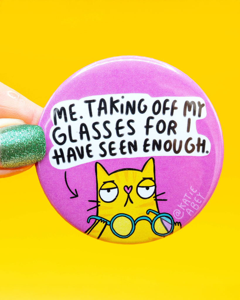 A round badge by Rebel Romance with a pink background, featuring a tired-looking yellow cat holding glasses with the text "Me. Taking off my glasses for I have seen enough." The badge is held up by a hand with sparkly green nail polish. A funny and relatable design by Katie Abey, perfect for bags, jackets, and gifts.