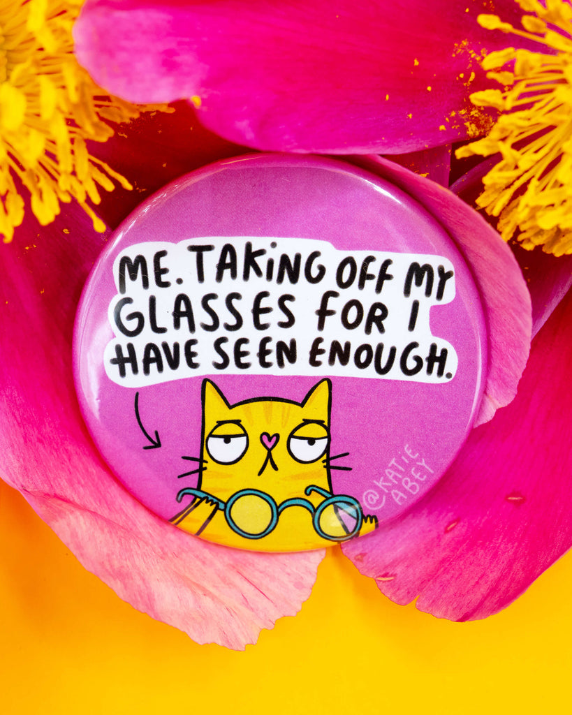 A round pink badge from Rebel Romance featuring a yellow cat with a grumpy expression removing its glasses. The badge says "Me. Taking off my glasses for I have seen enough." Designed by Katie Abey, the badge is surrounded by vibrant pink and yellow flower petals on a bold yellow background. A fun and quirky gift from an alternative UK small business.