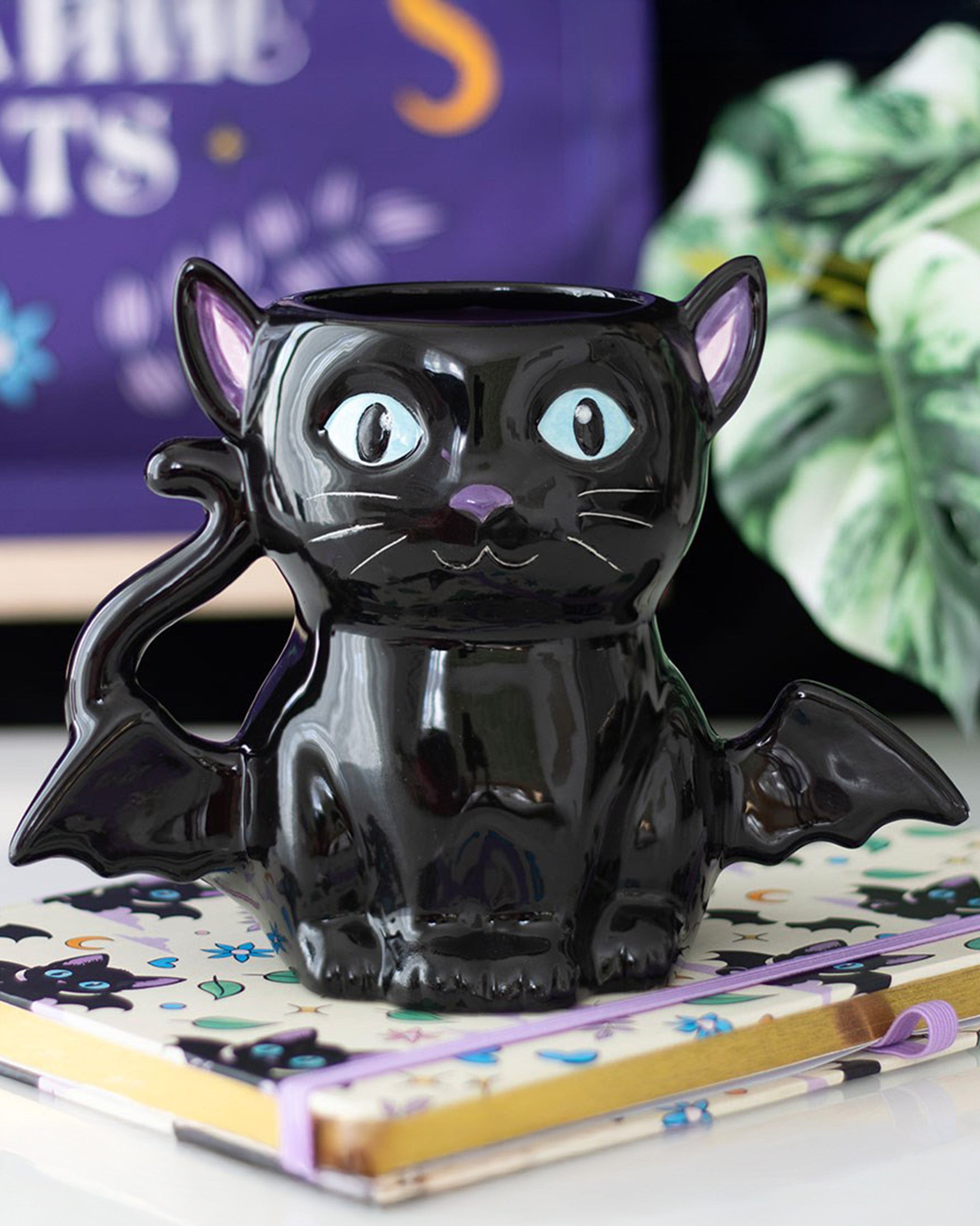 Black ceramic cat-shaped mug with bat wings, glossy finish, and purple accents on the ears, sitting on a cat-themed notebook.