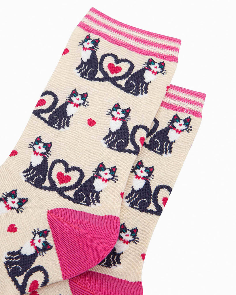 A pair of cream bamboo socks with a playful cat design, featuring black and white cats with their tails forming heart shapes, scattered pink hearts, and a striped pink cuff.