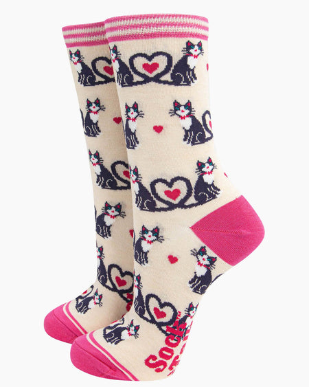 A pair of cream bamboo socks with a playful cat design, featuring black and white cats with their tails forming heart shapes, scattered pink hearts, and a striped pink cuff.