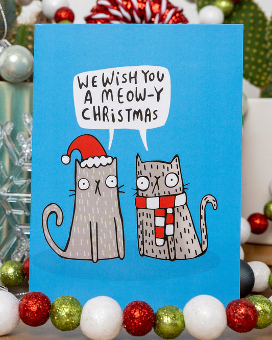 A colourful Christmas card from Rebel Romance featuring two cartoon cats on a bright blue background. One wears a red Santa hat and the other a red and white scarf. A speech bubble above them reads “We wish you a meow-y Christmas”. The card is surrounded by festive decorations including red, green, and white baubles. Designed by Katie Abey, this card is perfect for cat lovers and alternative Christmas vibes.