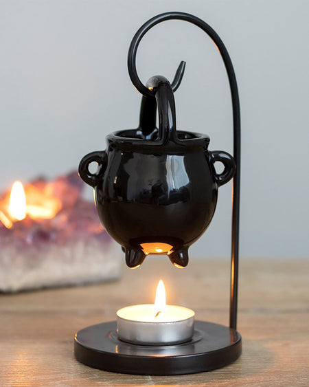 Hanging Cauldron Oil Burner by Rebel Romance. A glossy black ceramic cauldron suspended from a curved black metal stand, with a lit tealight beneath for warming wax melts or fragrance oils.
