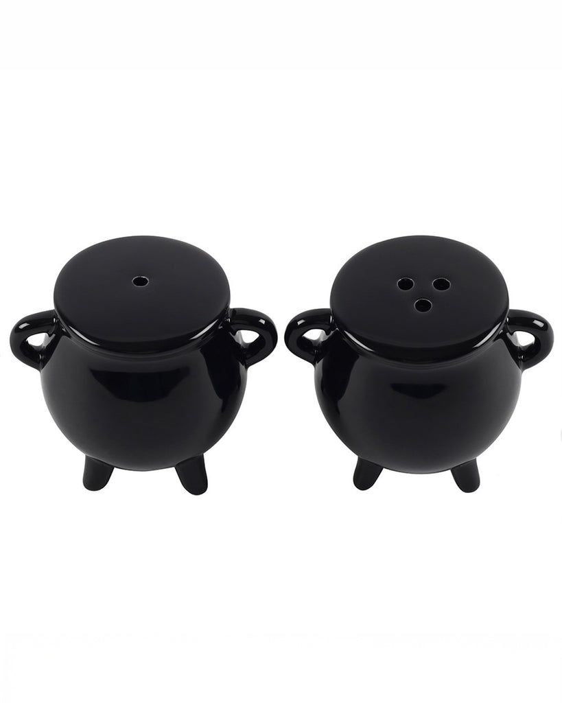 Studio shot of the Cauldron Salt & Pepper Shaker Set, featuring two glossy black ceramic shakers shaped like mini cauldrons. Each shaker has two side handles, three legs, and a rubber bottom stopper. One shaker has a single-hole lid, and the other has three holes. Ideal for adding witchy vibes to your kitchen decor.