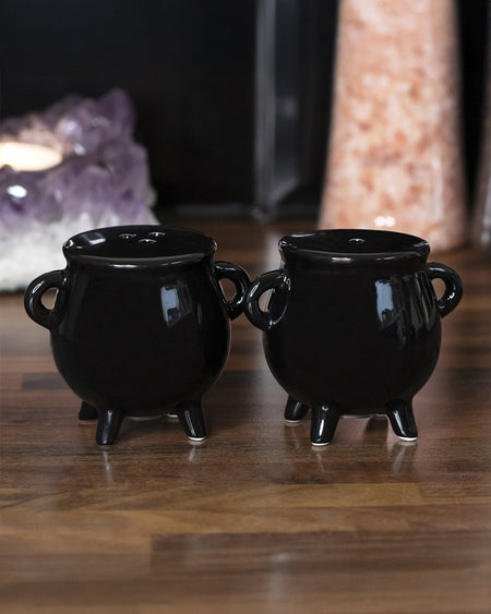Close-up of the Cauldron Salt & Pepper Shaker Set in glossy black ceramic. Both shakers have two side handles and stand on three short legs. One has a single-hole top, while the other has three holes for easy identification. Perfect for adding gothic charm to your kitchen. From Rebel Romance, a small UK brand embracing alternative style.