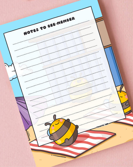 "Notes to Bee-member" A6 notepad by Toastedink, available at Rebel Romance, featuring a cute cartoon bee relaxing on a red and white striped towel. Set on a pastel pink background.