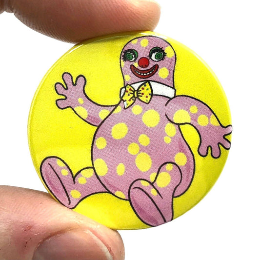 Fun and quirky button badge from Rebel Romance held between fingers, showcasing a cheerful pink and yellow spotted character with a red clown nose and bow tie on a yellow background. Designed by Bite Your Granny, this nostalgic pin is perfect for adding eccentric flair to any alternative outfit or tote bag.