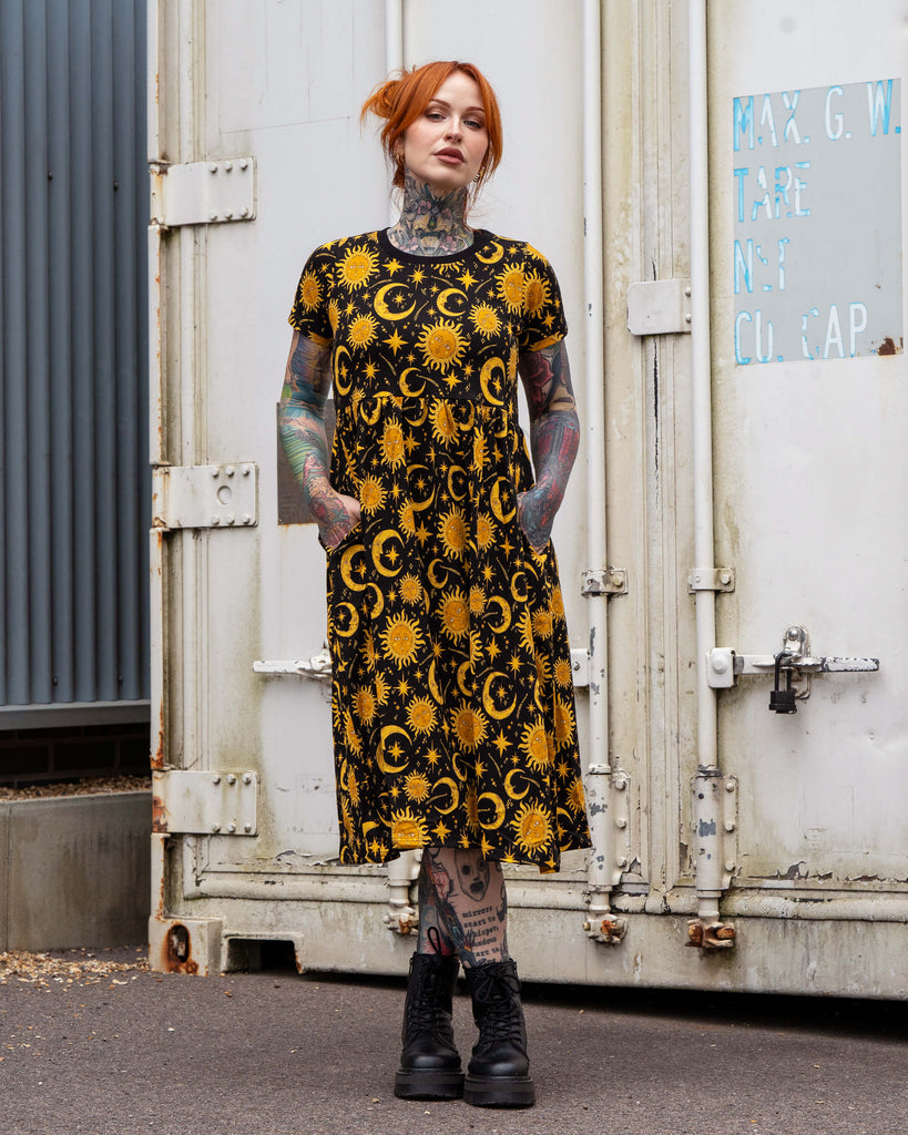 Erin wearing the Rebel Romance Celestial Midi Dress made from stretch cotton. The black dress features a bold yellow celestial print with suns, moons, and stars, short sleeves, and a relaxed fit. Styled with chunky black boots, this alternative fashion piece combines comfort and cosmic vibes.