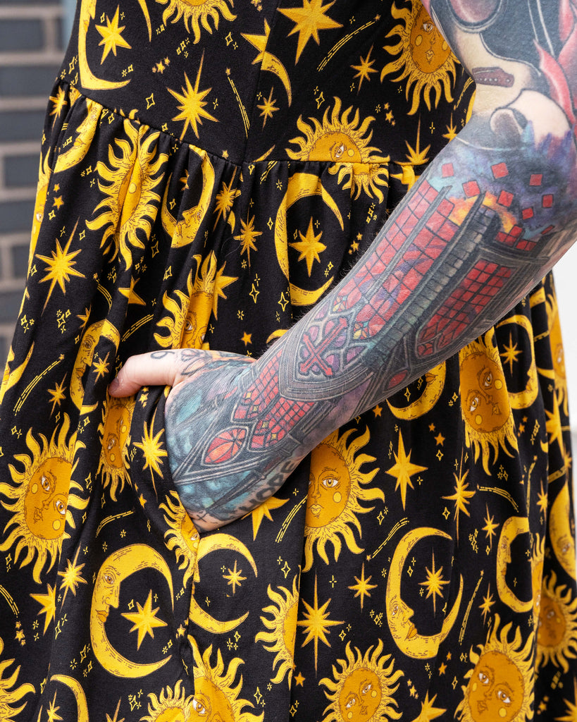 Detail shot of the Rebel Romance Celestial Midi Dress pocket, showing Erin’s tattooed arm resting inside. The stretch cotton fabric is covered in a golden celestial print of suns, moons, and stars, perfect for alternative fashion lovers.