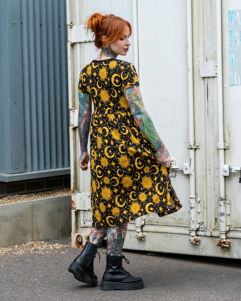 Back view of Erin modelling the Rebel Romance Celestial Midi Dress in stretch cotton. The black smock dress is printed with yellow suns, crescent moons, and stars for a celestial, witchy-inspired alternative fashion look.