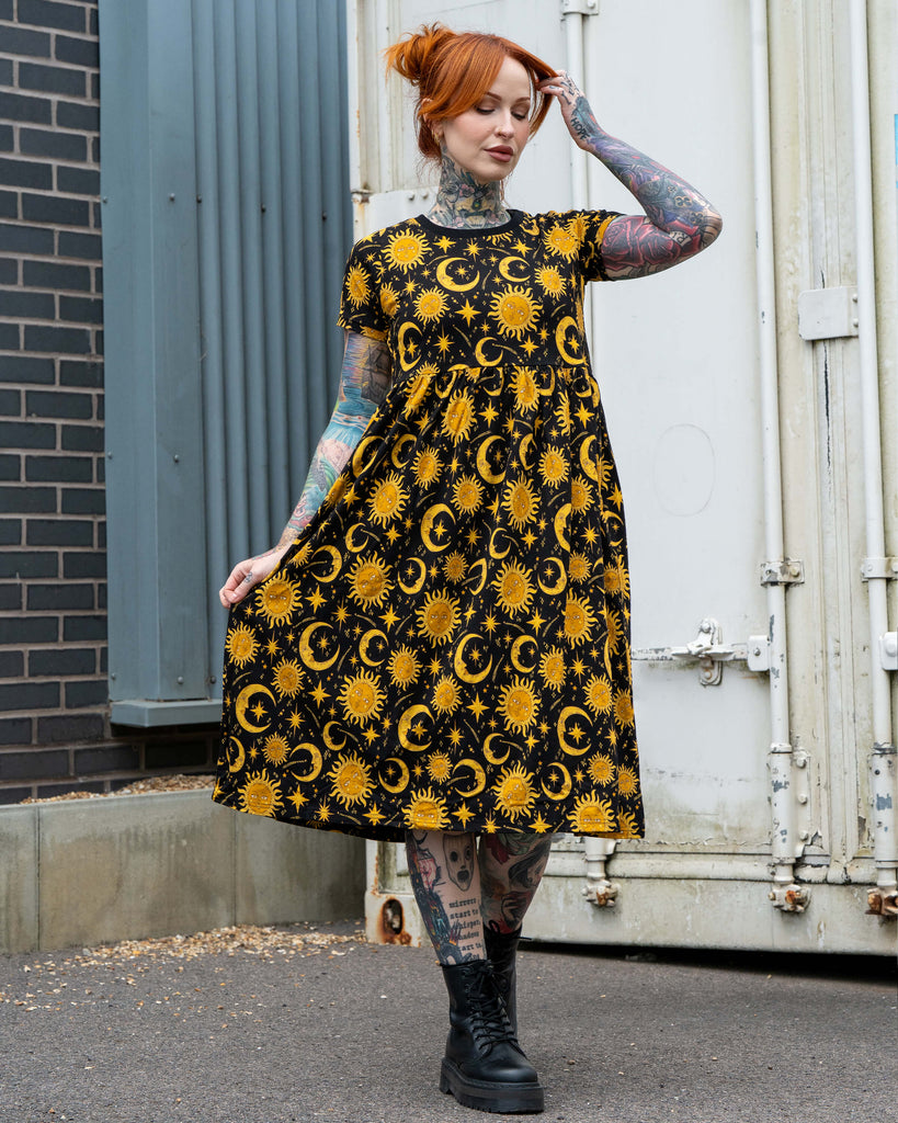 Erin wearing the Rebel Romance Celestial Midi Dress, styled with chunky boots. The stretch cotton dress features a relaxed fit, short sleeves, pockets, and a yellow celestial print of suns and moons on black, making it perfect for everyday alternative fashion.