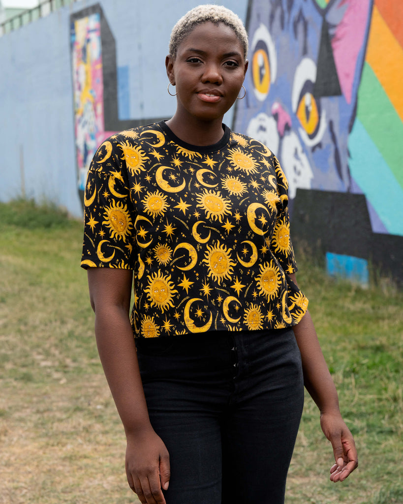 Chish wears the Rebel Romance Celestial Tee with black jeans. The cropped T-shirt features a golden yellow sun, moon and star print on black, styled against a street art mural with a painted cat face.
