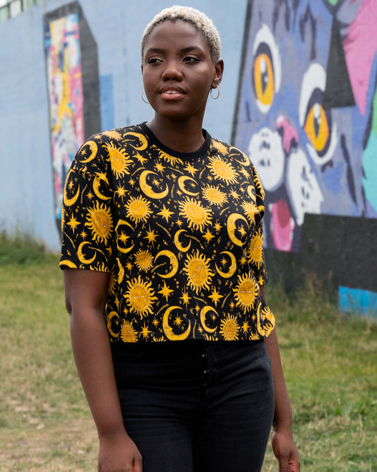 Chish wears the Celestial Tee by Rebel Romance, styled with black high-waisted jeans. The unisex crop T-shirt has an all-over golden celestial print with suns, moons and stars on black.