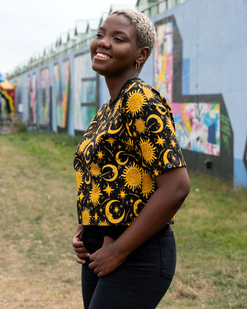 Chish models the Rebel Romance Celestial Tee in a side profile pose, smiling. The black crop top is covered in a golden celestial pattern of suns, moons and stars, styled with black jeans for a mystical yet casual look.