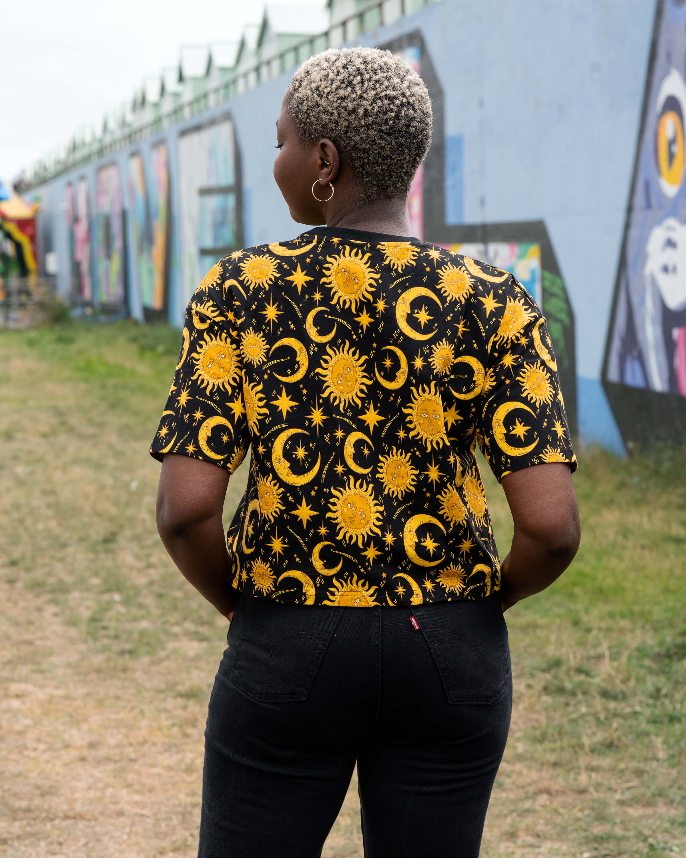 Back view of Chish wearing the Rebel Romance Celestial Tee. The black cropped T-shirt shows a repeat pattern of golden suns, crescent moons and stars, styled with black jeans in an outdoor streetwear shoot.