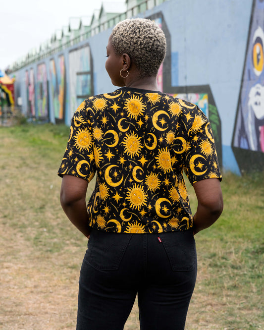 Back view of Chish wearing the Rebel Romance Celestial Tee. The black cropped T-shirt shows a repeat pattern of golden suns, crescent moons and stars, styled with black jeans in an outdoor streetwear shoot.