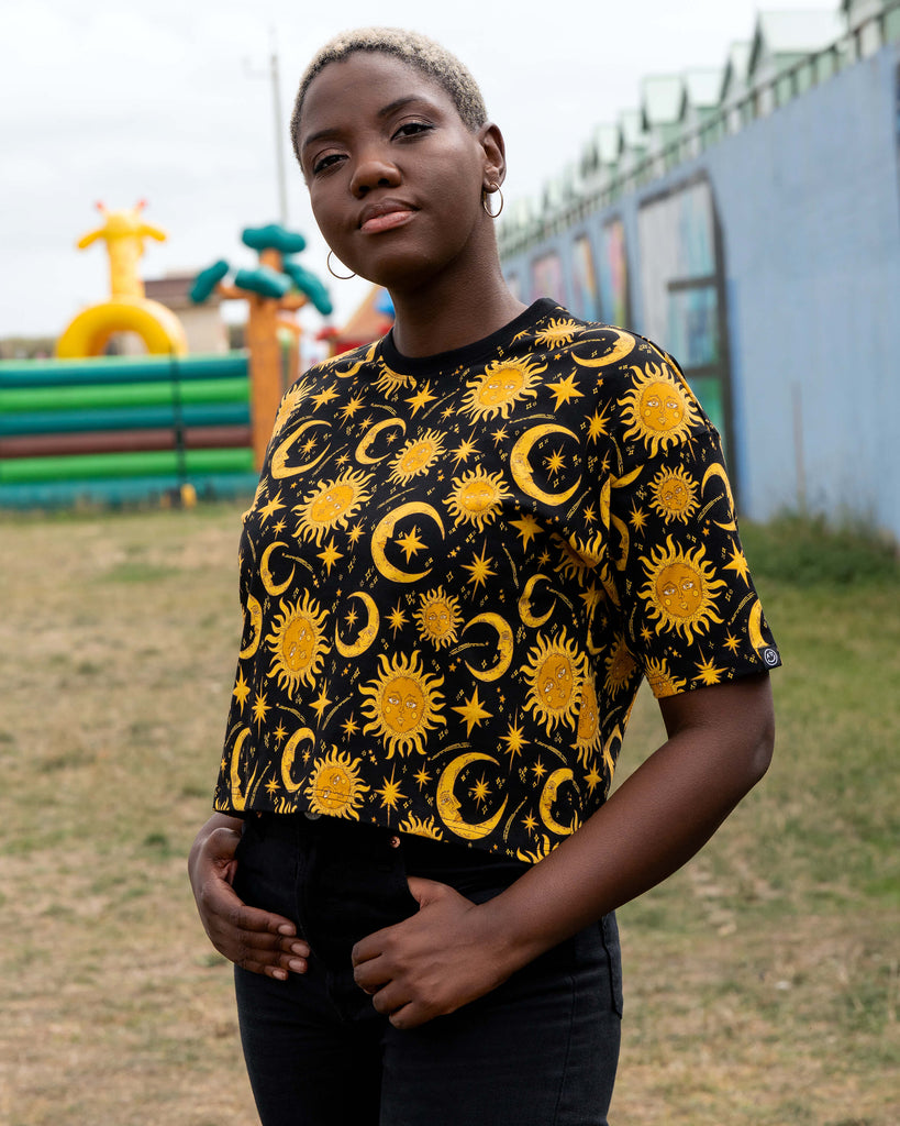 Chish models the Rebel Romance Celestial Tee, styled with black jeans. The celestial crop top features a mystical golden sun and moon print on black, photographed outdoors with colourful playground inflatables in the background.
