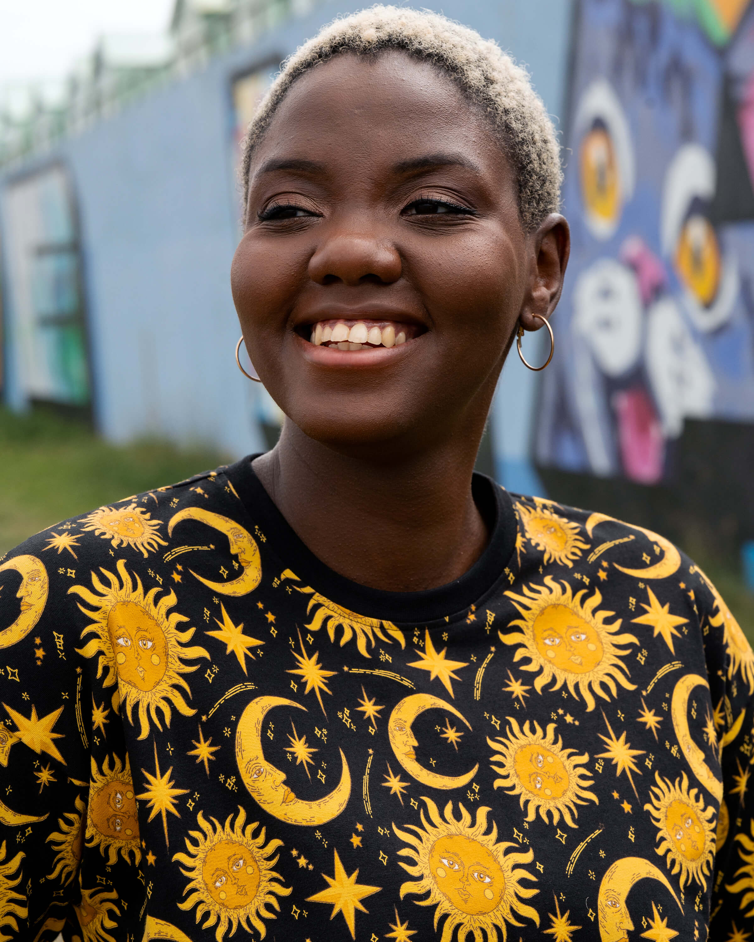 Portrait of Chish smiling in the Rebel Romance Celestial Tee. The black T-shirt features a vibrant golden celestial print with suns, moons and stars, styled with hoop earrings for a witchy alternative look.