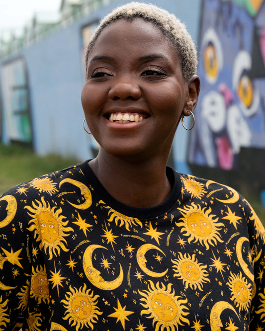Portrait of Chish smiling in the Rebel Romance Celestial Tee. The black T-shirt features a vibrant golden celestial print with suns, moons and stars, styled with hoop earrings for a witchy alternative look.