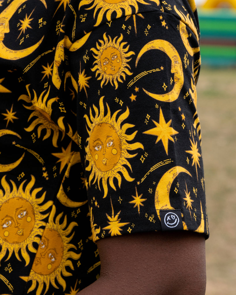 Close-up of the Rebel Romance Celestial Tee worn by Chish, showing the black fabric patterned with golden suns, moons and stars. A small Rebel Romance smiley logo tag is stitched on the sleeve hem.