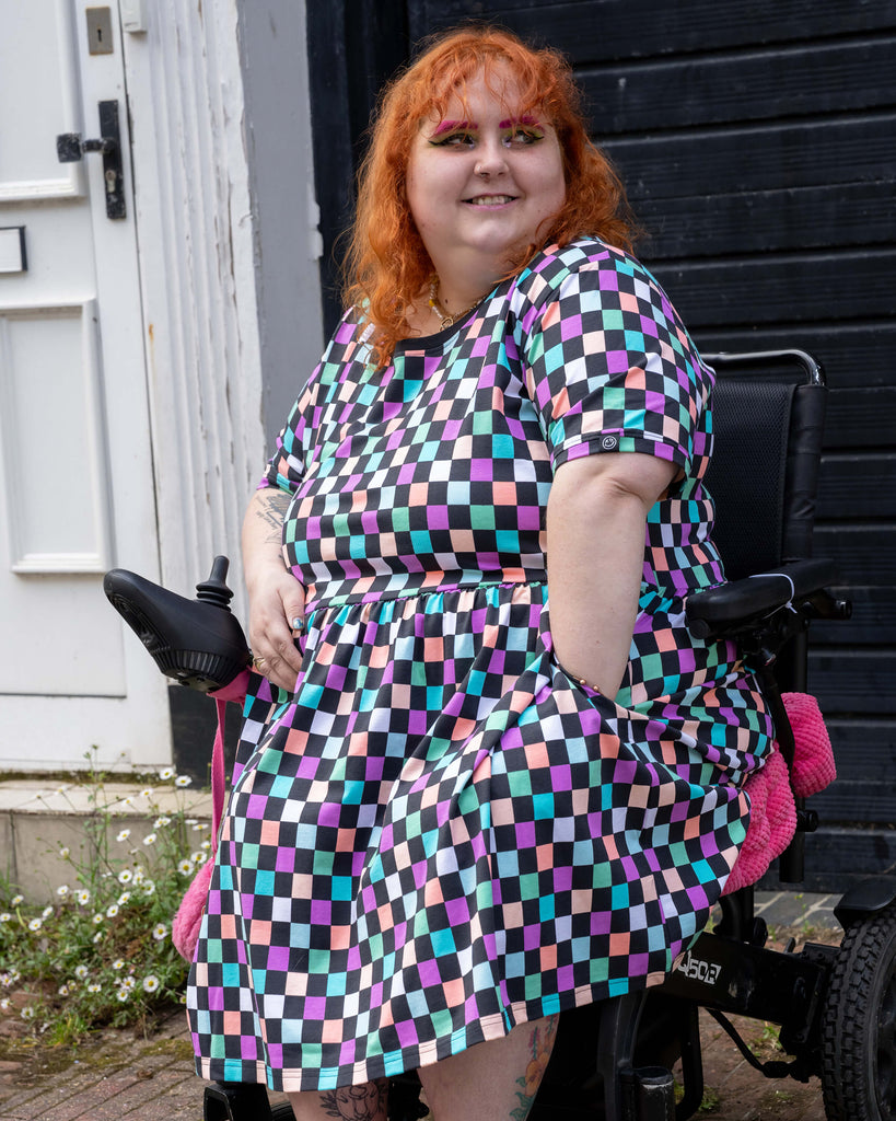 Lauren sits in a power wheelchair, smiling while wearing the Rebel Romance pastel checkerboard stretch smock dress in size 24. The dress features a colourful pattern with pink, teal, purple, and black squares, short sleeves, and pockets. Styled with bright pink Crocs and bold eye makeup, Lauren poses outside against a black wooden door and white wall.