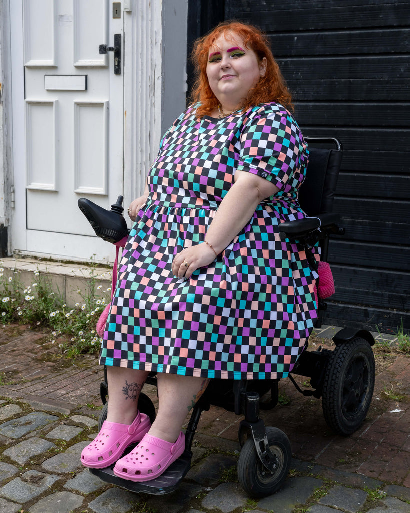 Lauren models the Rebel Romance checkerboard smock dress while seated in a power wheelchair on a cobbled street. The dress has a gathered waist, short sleeves, and a playful multicoloured square print. She completes the look with vibrant pink Crocs and colourful makeup, exuding joyful energy in front of a black and white building exterior.