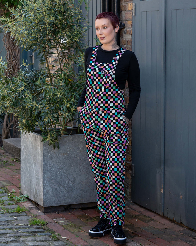 Katie, an alternative tattooed woman wearing a colourful checkerboard patterned dungarees in colour block colours of blue, orange, purple, green, blue and pastels. They've paired it with black trainers and a black long sleeve tshirt underneath standing outside looking left with their hands in their pockets in front of a grey door.