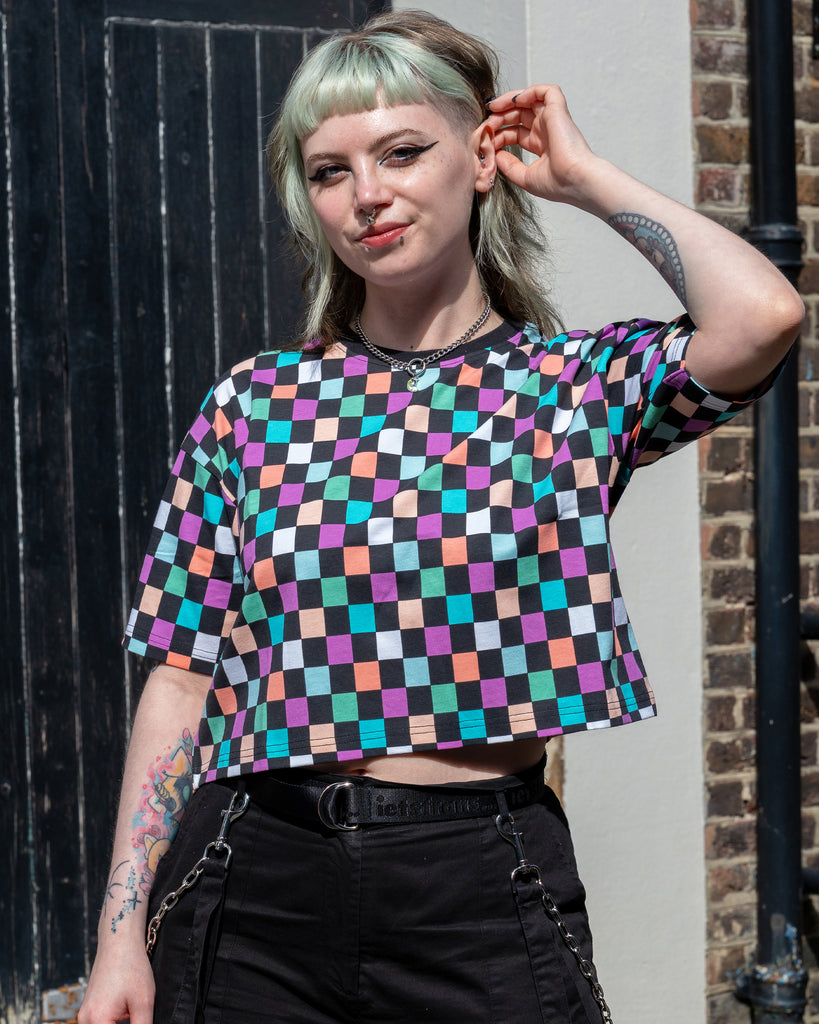 Faeryn poses with one hand raised, showing the full front of the Rebel Romance Checkerboard Tee in pastel pink, lilac, green, peach and turquoise tones. Styled with high-waisted black trousers and silver accessories.