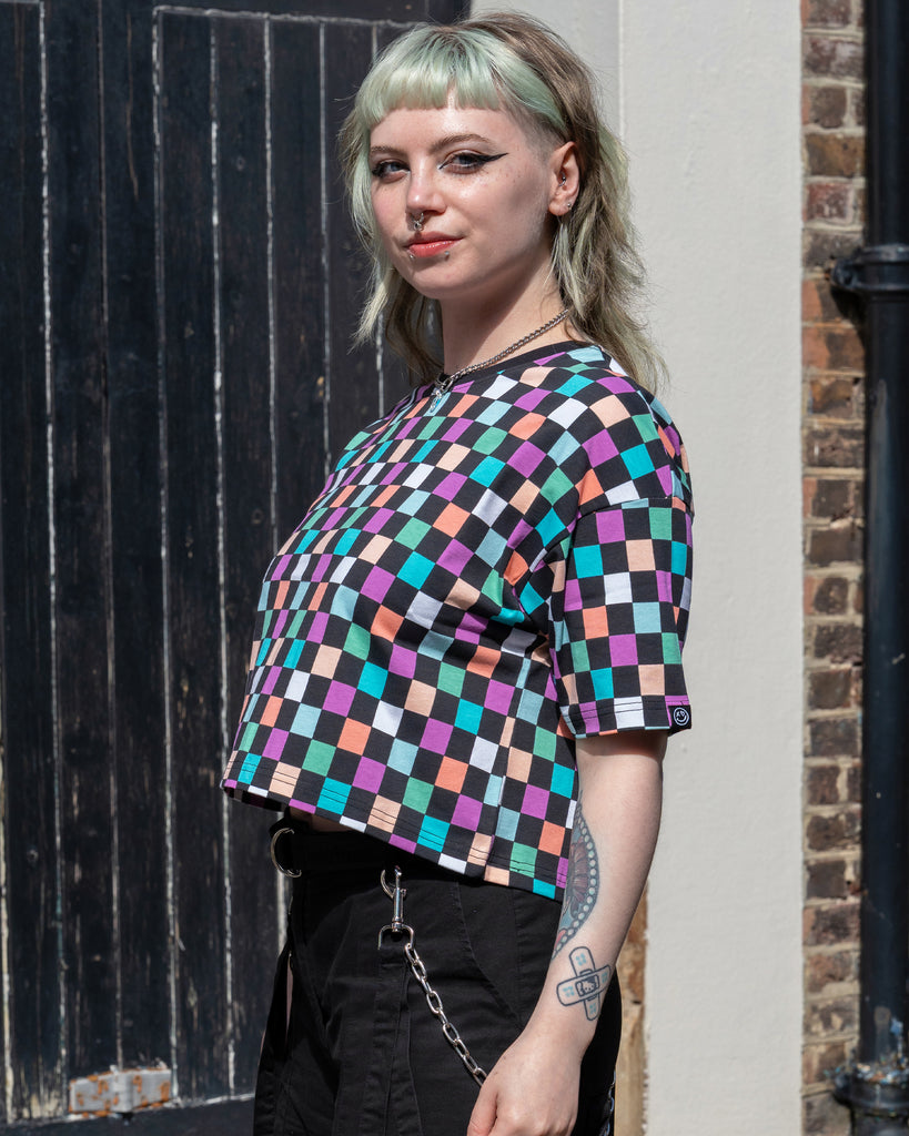 Faeryn stands side-on in bright sunlight, showing the cropped fit of the Rebel Romance Checkerboard Boxy Tee. The multicolour check print contrasts with their black trousers and visible arm tattoos.