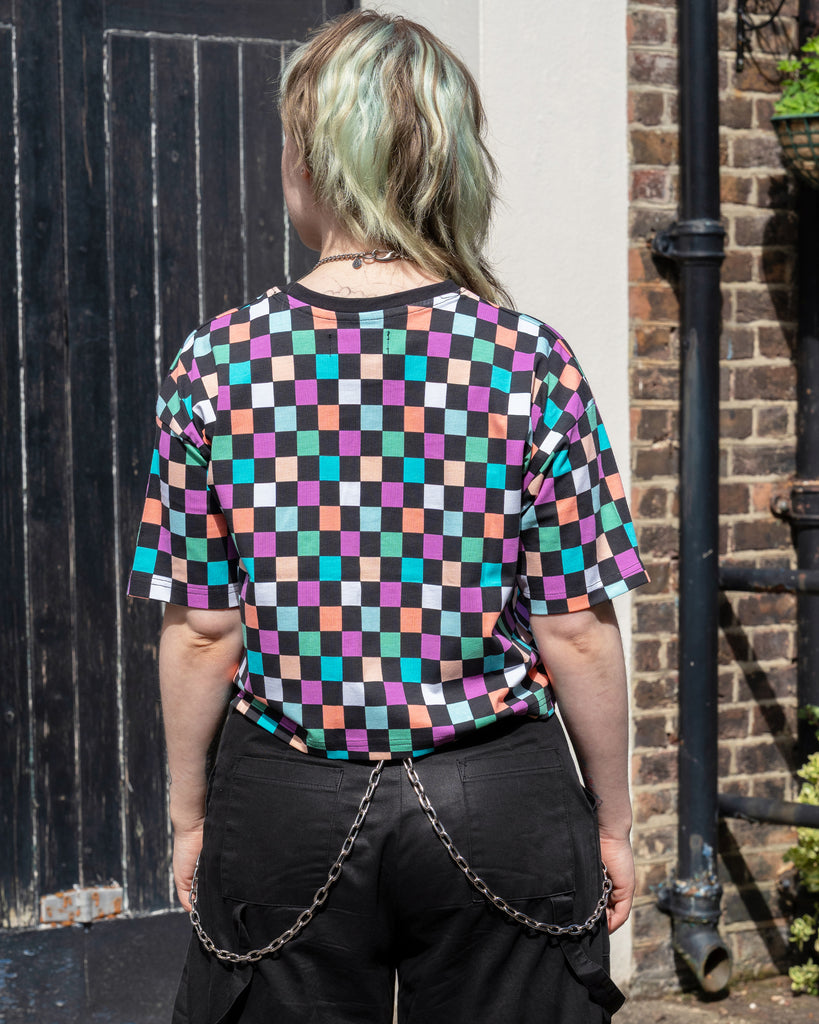Back view of model Faeryn wearing the Rebel Romance Checkerboard Boxy Tee. The vibrant pastel check pattern covers the back and sleeves, styled with black trousers and a silver chain belt.
