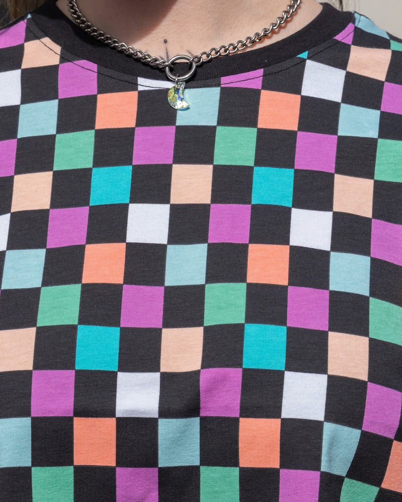 Zoomed-in shot of the Rebel Romance Checkerboard Tee's chest area, showing the soft pastel squares and black neckline paired with a chunky silver moon charm necklace worn by model Faeryn.