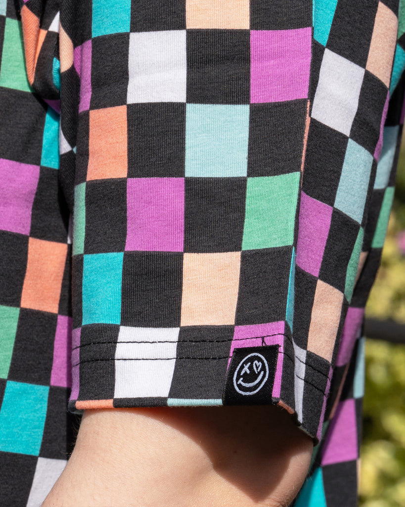 Close-up of the Rebel Romance Checkerboard Boxy Tee sleeve, showing a pastel check pattern and the black woven label with the Rebel Romance smiley face logo. The colourful print includes pink, peach, mint, teal, and lilac blocks.