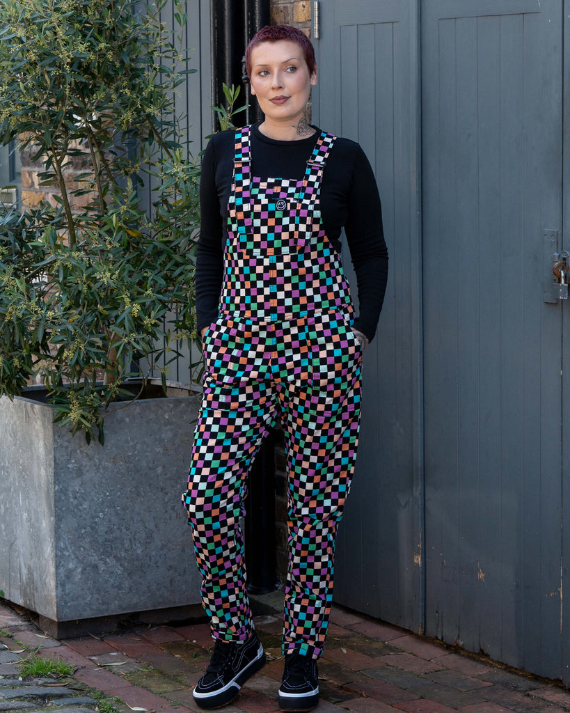 Katie, a tattooed woman wearing a colourful checkerboard patterned dungarees in colour block colours of blue, orange, purple, green, blue and pastels. She's paired it with black trainers and a black long sleeve tshirt underneath standing in front of a grey door. She's got her hands in her pockets looking right. 
