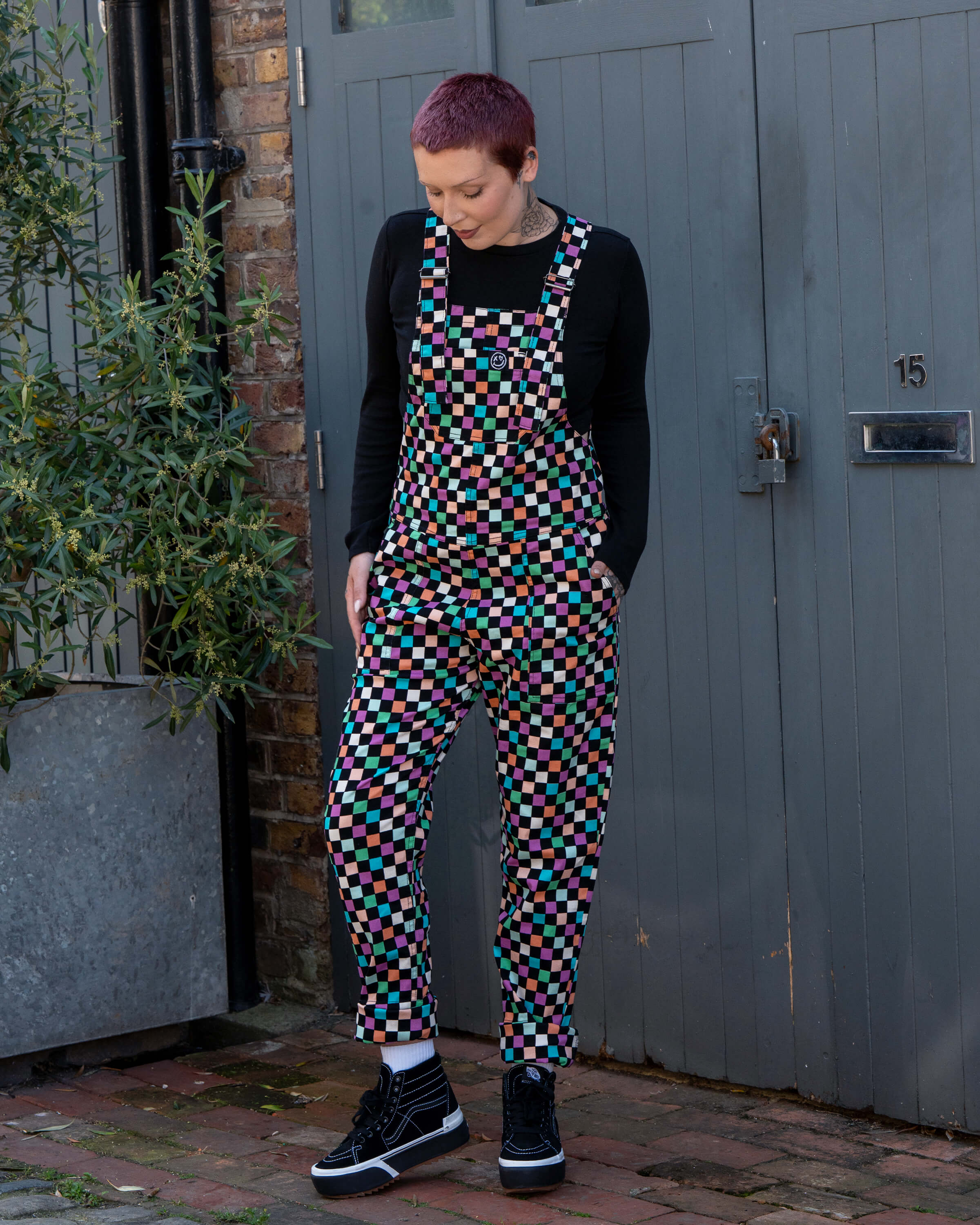 Katie, a tattooed woman wearing a colourful checkerboard patterned dungarees in colour block colours of blue, orange, purple, green, blue and pastels. They've paired it with black trainers and a black long sleeve tshirt underneath standing in front of a grey door. She is facing forward looking down at the dungarees with both hands in her pockets. 
