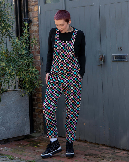Katie, a tattooed woman wearing a colourful checkerboard patterned dungarees in colour block colours of blue, orange, purple, green, blue and pastels. They've paired it with black trainers and a black long sleeve tshirt underneath standing in front of a grey door. She is facing forward looking down at the dungarees with both hands in her pockets. 