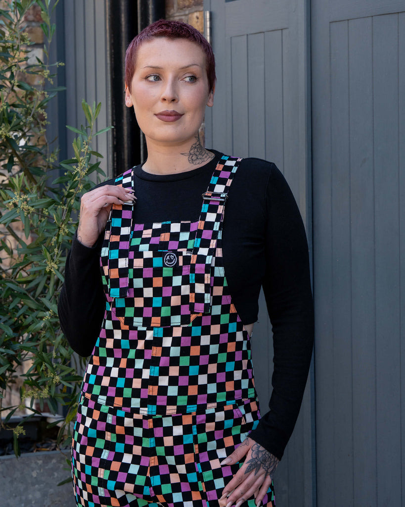 Close up of Katie, a tattooed woman wearing a colourful checkerboard patterned dungarees in colour block colours of blue, orange, purple, green, blue and pastels. They've paired it with a black long sleeve tshirt underneath standing in front of a grey door.