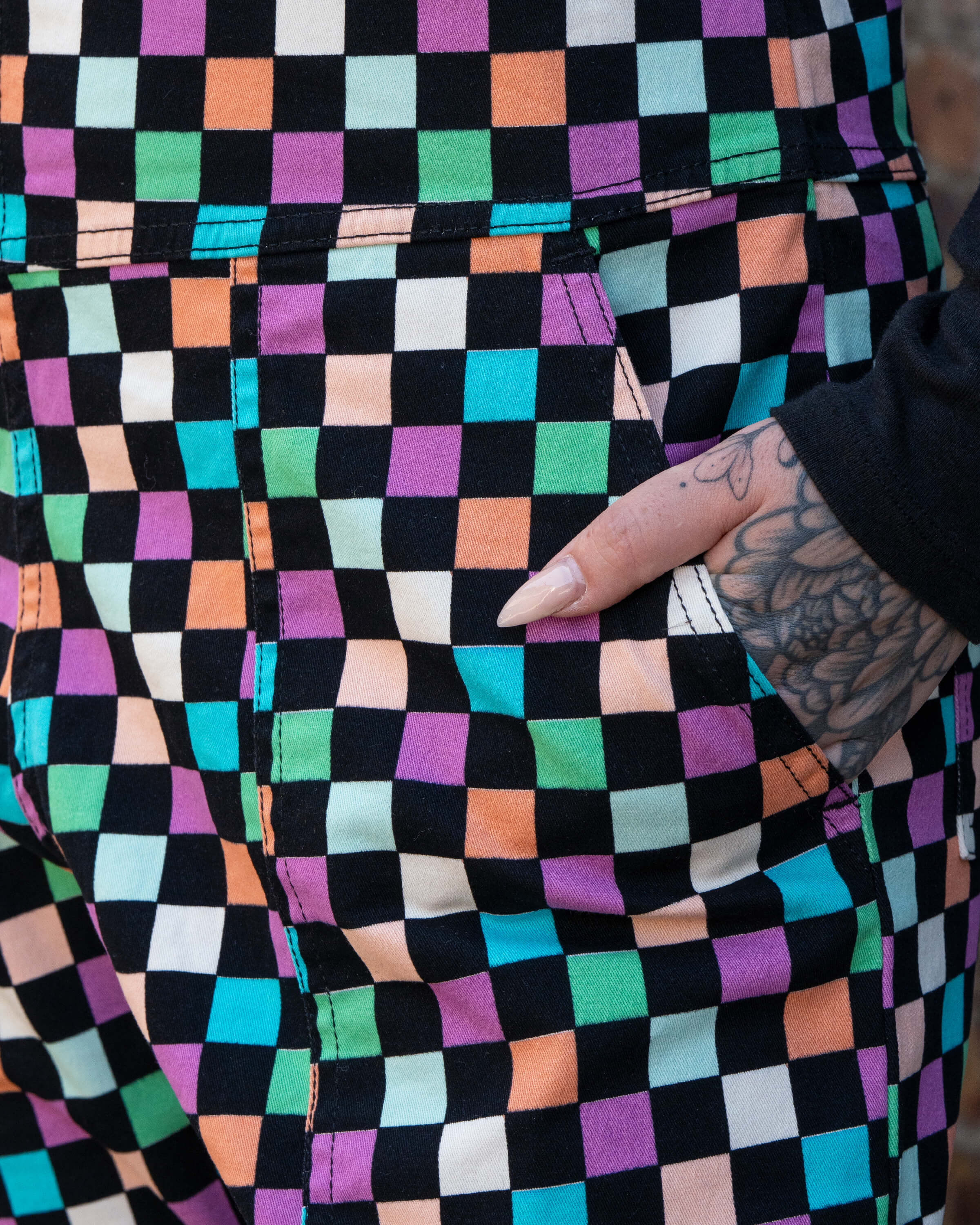 Close up of Katie, a tattooed woman wearing a colourful checkerboard patterned dungarees in colour block colours of blue, orange, purple, green, blue and pastels. They've paired it with a black long sleeve tshirt underneath with their hand in the pocket. 