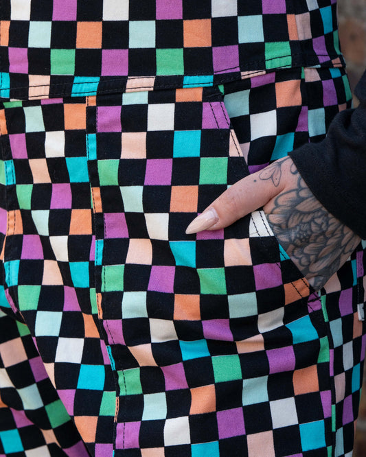 Close up of Katie, a tattooed woman wearing a colourful checkerboard patterned dungarees in colour block colours of blue, orange, purple, green, blue and pastels. They've paired it with a black long sleeve tshirt underneath with their hand in the pocket. 