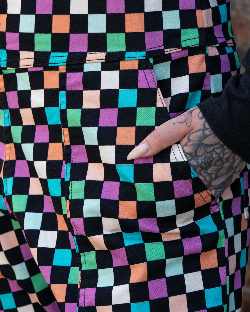 Close up of Katie, a tattooed woman wearing a colourful checkerboard patterned dungarees in colour block colours of blue, orange, purple, green, blue and pastels. They've paired it with a black long sleeve tshirt underneath with their hand in the pocket. 