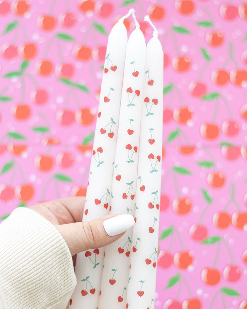 The Rebel Romance Heart Cherry Taper Candles set displayed on a pink background with matching cherry print paper. The packaging's cute red and pink design complements the candles' playful heart-shaped cherry pattern.