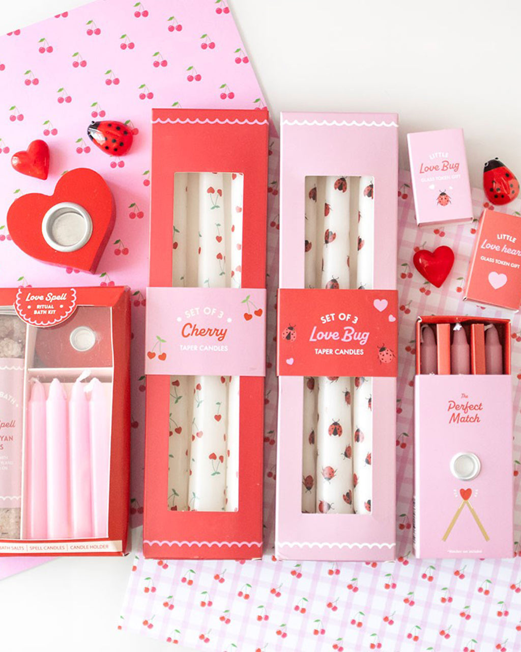 A vibrant flat lay of Valentine's-themed homeware and gifts, including the Rebel Romance Heart Cherry Taper Candles set. The red and pink packaging showcases the three white candles, each adorned with an adorable cherry heart pattern. Perfect for gifting or adding a cute touch to a romantic table setting.